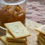 A jar of chutney and some almond parmesan crackers on a wooden serving board