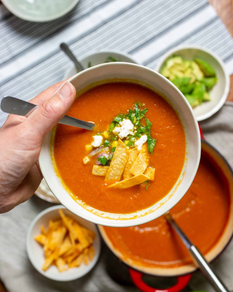 a hand holding a bowl of tortilla soup with a pot of soup and fixins in the background.