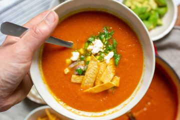 a hand holding a bowl of tortilla soup with a pot of soup and fixins in the background.