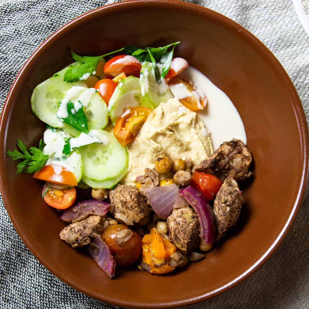 Shawarma spiced chicken cubes with hummus, cucumber and tomato salad, and yogurt sauce in a brown bowl on a linen napkin.