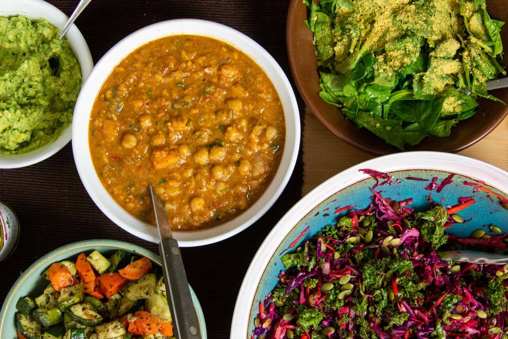 Dishes filled with toppings at a baked potato party. Moroccan chickpea stew, winter slaw, spinach salad, and guacamole.