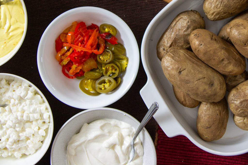 A pile of baked potatoes next to some small bowls of pickled peppers, sour cream, and cottage cheese
