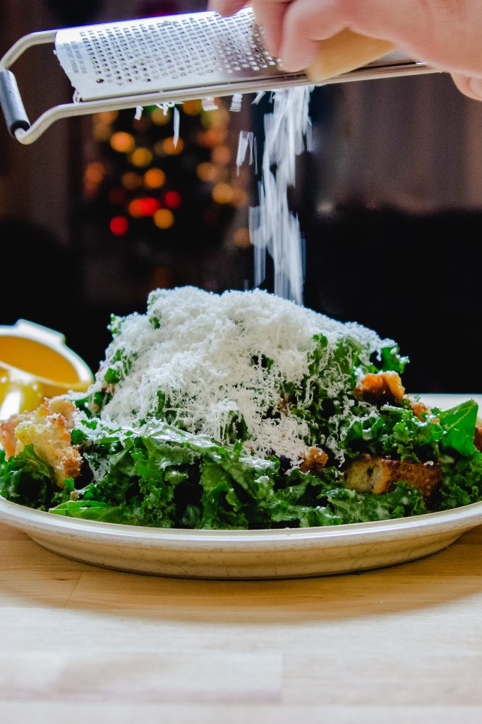 a plate of kale caesar salad with someone grating parmesan on top.