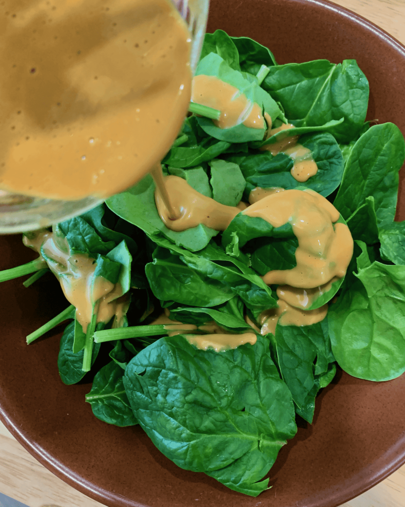 Pouring nutritional yeast dressing over baby spinach in a bowl.