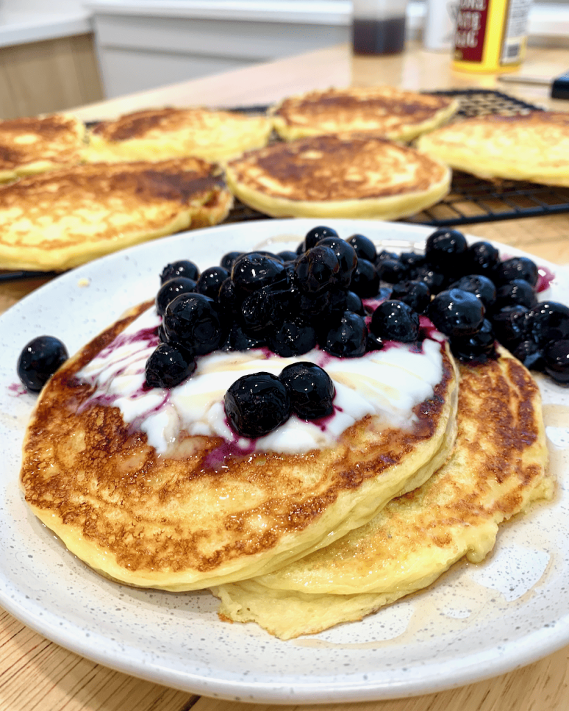 Protein pancakes with yogurt and blueberries on a plate, with cooling pancakes in the background. 