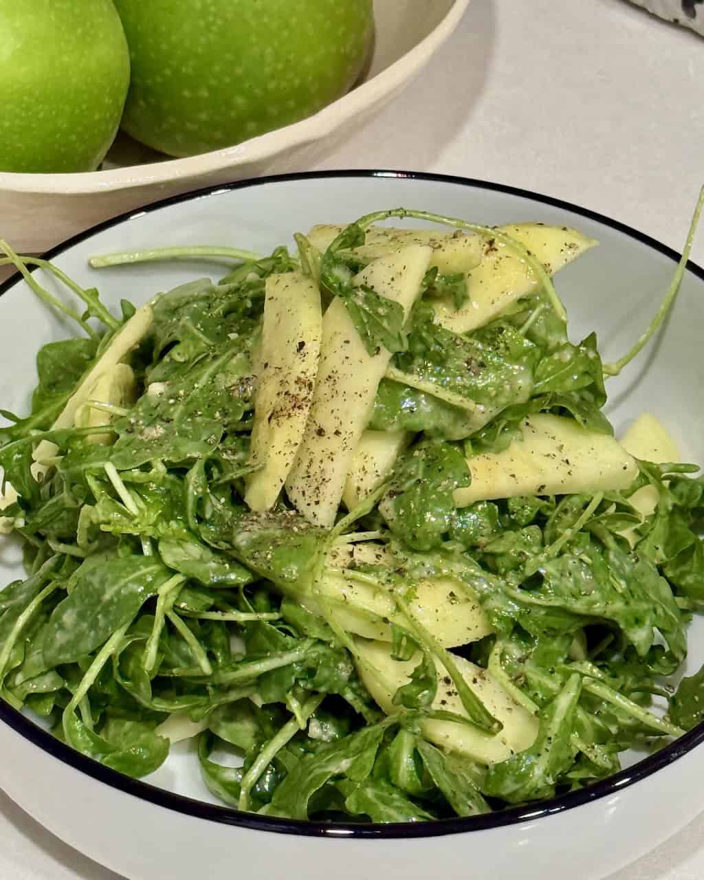 Arugula salad in a serving bowl for one. Green apples in the background.