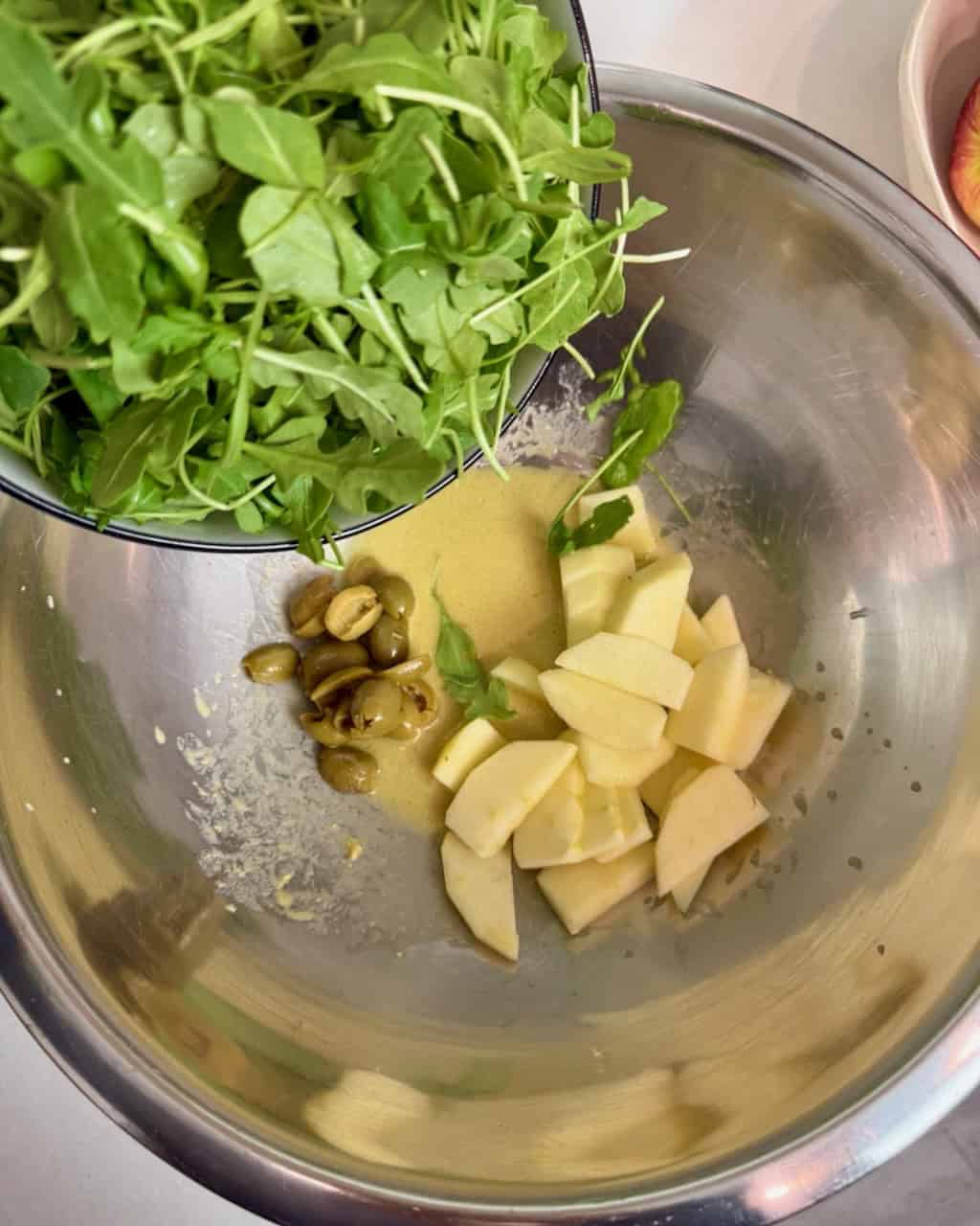 Adding fresh arugula to the sliced apples, olives, and dressing, in a bowl.