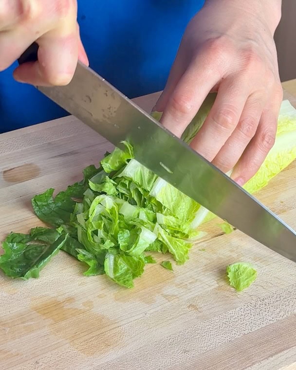 A woman slices romaine lettuce on a wooden cutting board.