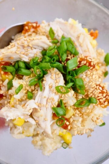 A bowl of chicken, rice, and corn, garnished with sliced green onions, toasted sesame seeds, and hot sauce.