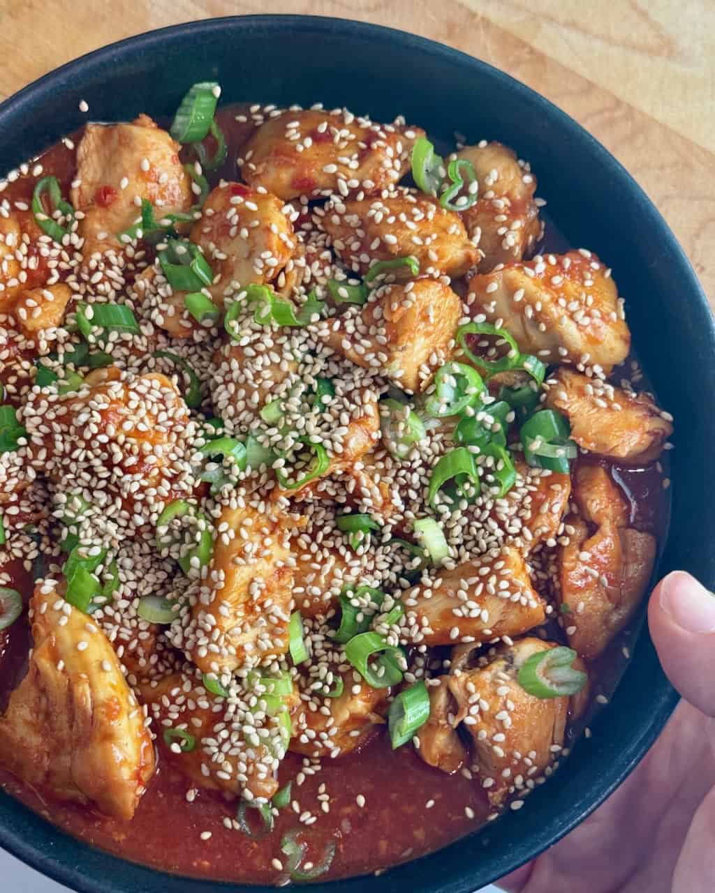 A woman holds a plate of gochujang chicken with sesame seeds and green onions.