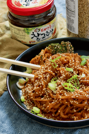 A plate of gochujang noodles and chopsticks with a jar of gochujang and a jar of sesame seeds on the side.