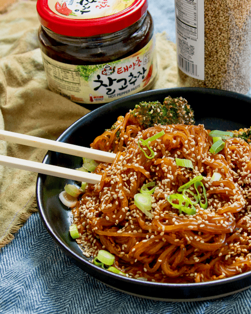 A plate of gochujang noodles and chopsticks with a jar of gochujang and a jar of sesame seeds on the side.