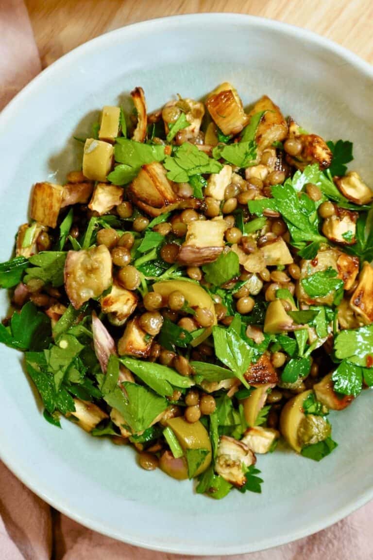 A bowl of eggplant lentil salad with a pink napkin on a wooden countertop.