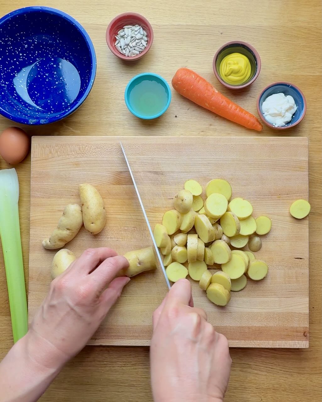 A woman slices fingerling potatoes on a wooden cutting board with potato salad ingredients around.