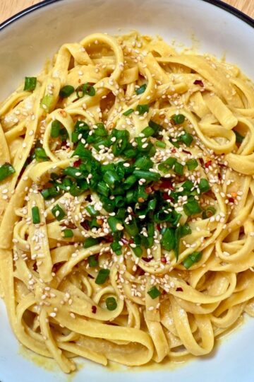 Nutritional yeast noodles garnished with green onions and sesame seeds in a white bowl.