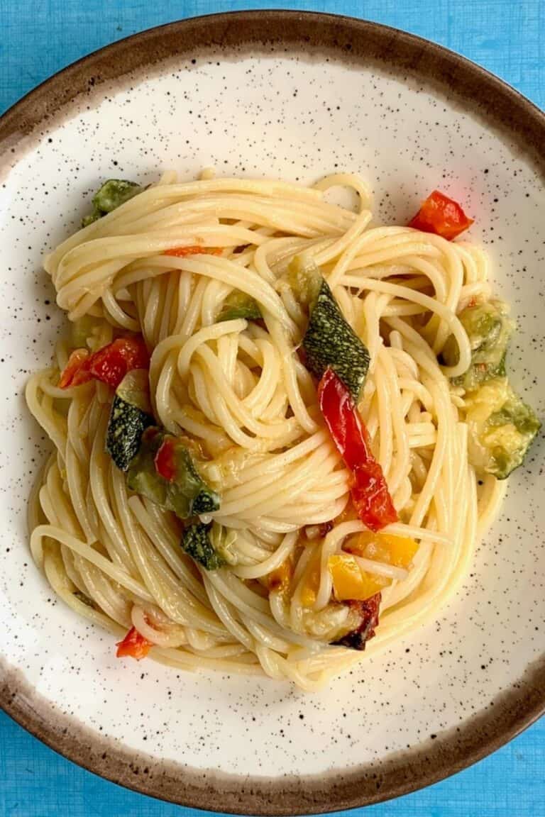 Spaghetti tossed with zucchini and pepper sauce with parmesan cheese sprinkled on top. The spaghetti is on a brown plate on a blue table.