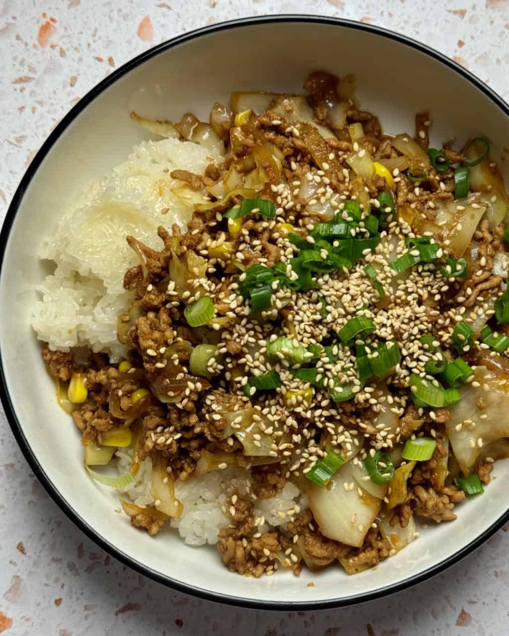 Overhead image of pork and cabbage stir fry over rice in a white bowl with black trim.