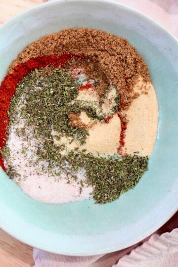 Herbs, spices, and salt, partially mixed together in a green bowl on a pink tea towel.