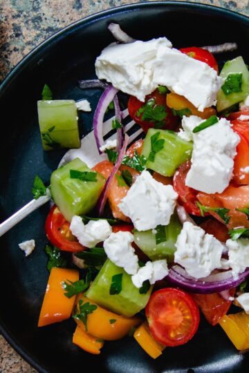 Bulgarian Shopska Salad in a blue bowl with some red onion an cherry tomatoes on the side.