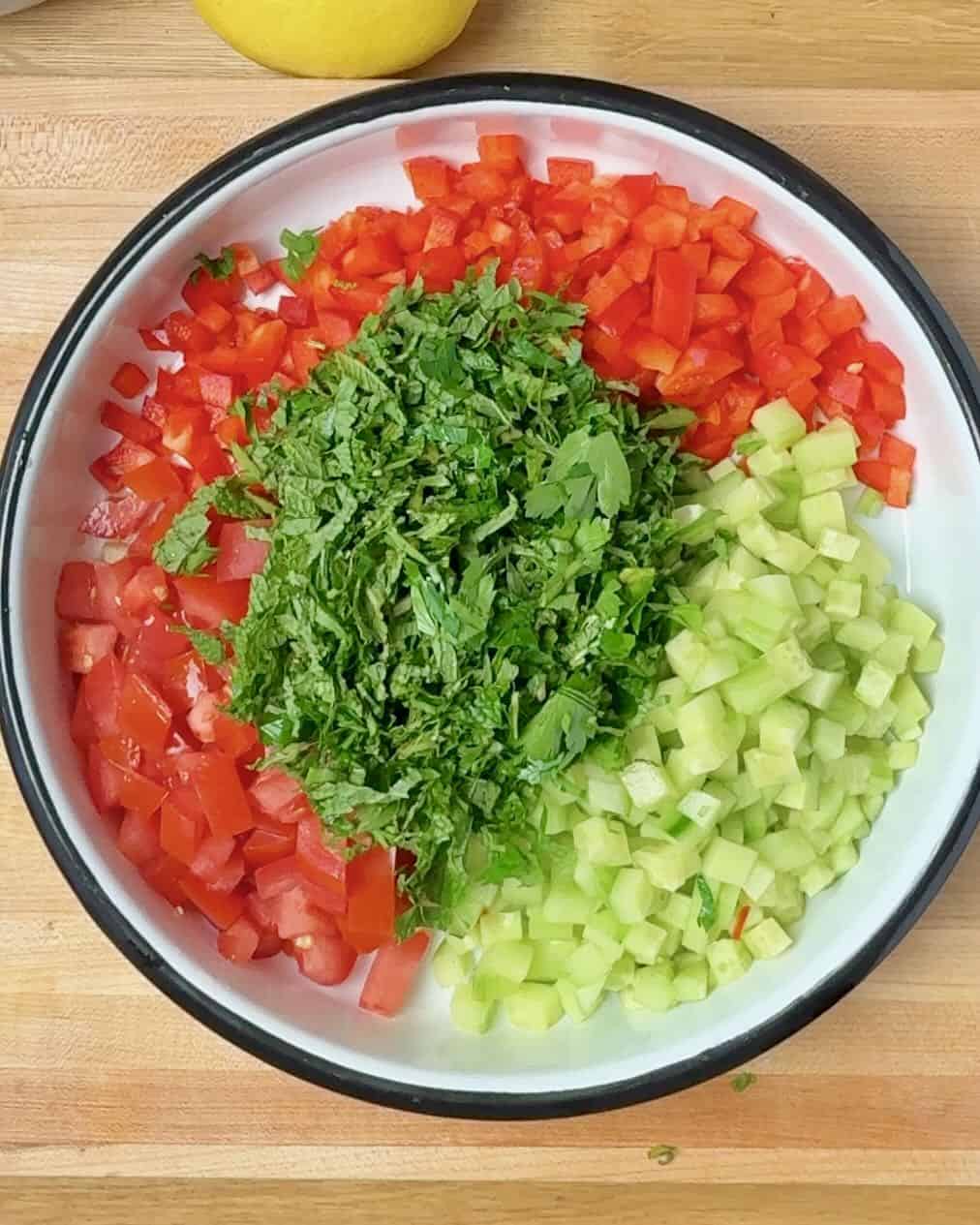 Chopped tomatoes, cucumbers, red bell peppers, mint and parsley in a bowl.