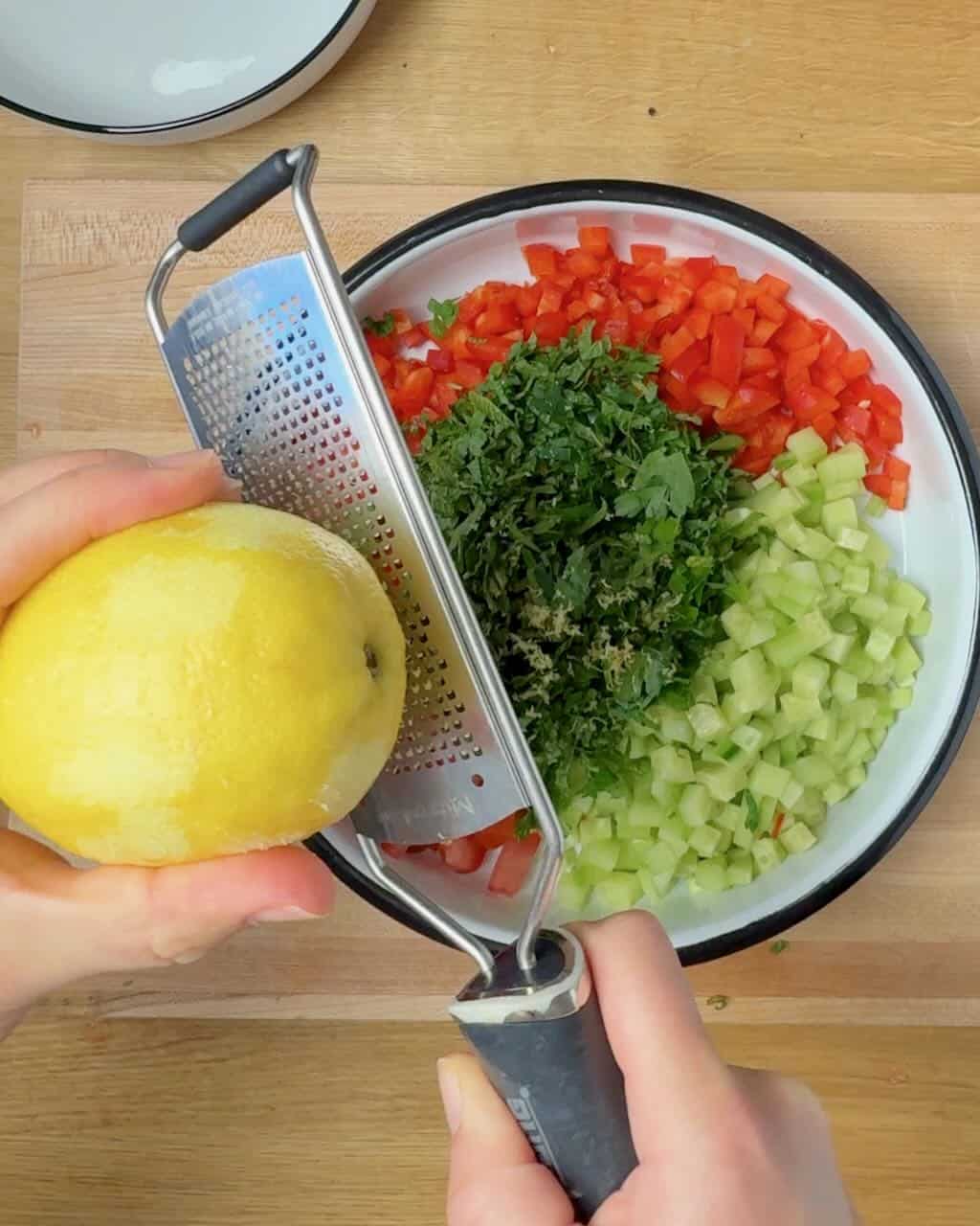 A woman grates lemon zest into a bowl of chopped vegetables and herbs.