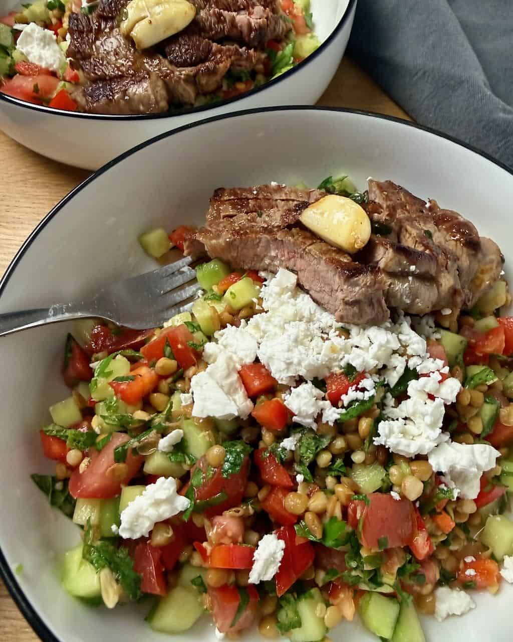 Grilled steak with fresh vegetable salad and feta cheese in a white bowl.