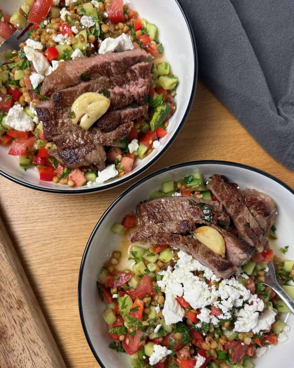 Juicy grilled steak slices with fresh lentil salad and crumbled feta cheese in white bowls.