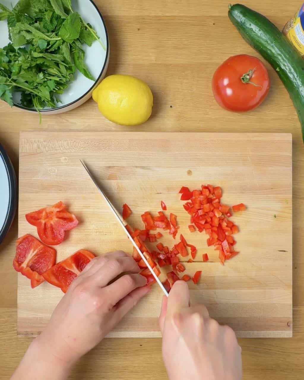 A woman chops red bell pepper on a wooden cutting board with fresh vegetables around it.
