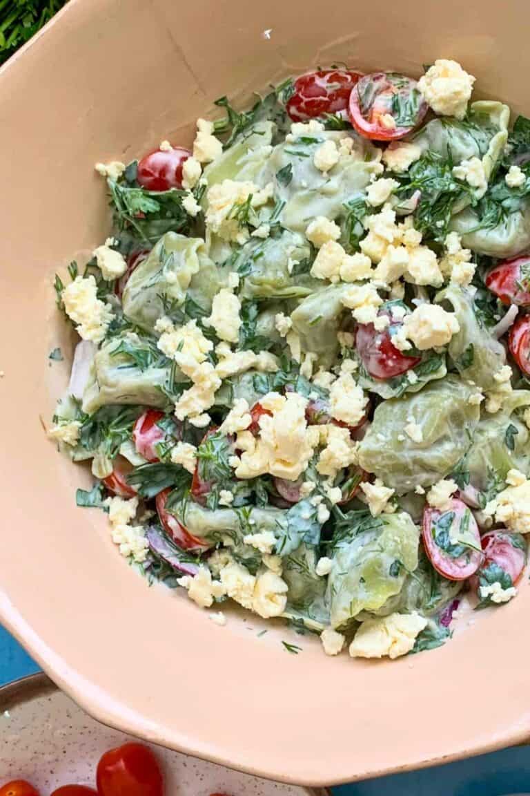 A large bowl of tortellini salad with a bowl of cherry tomatoes on a blue table.