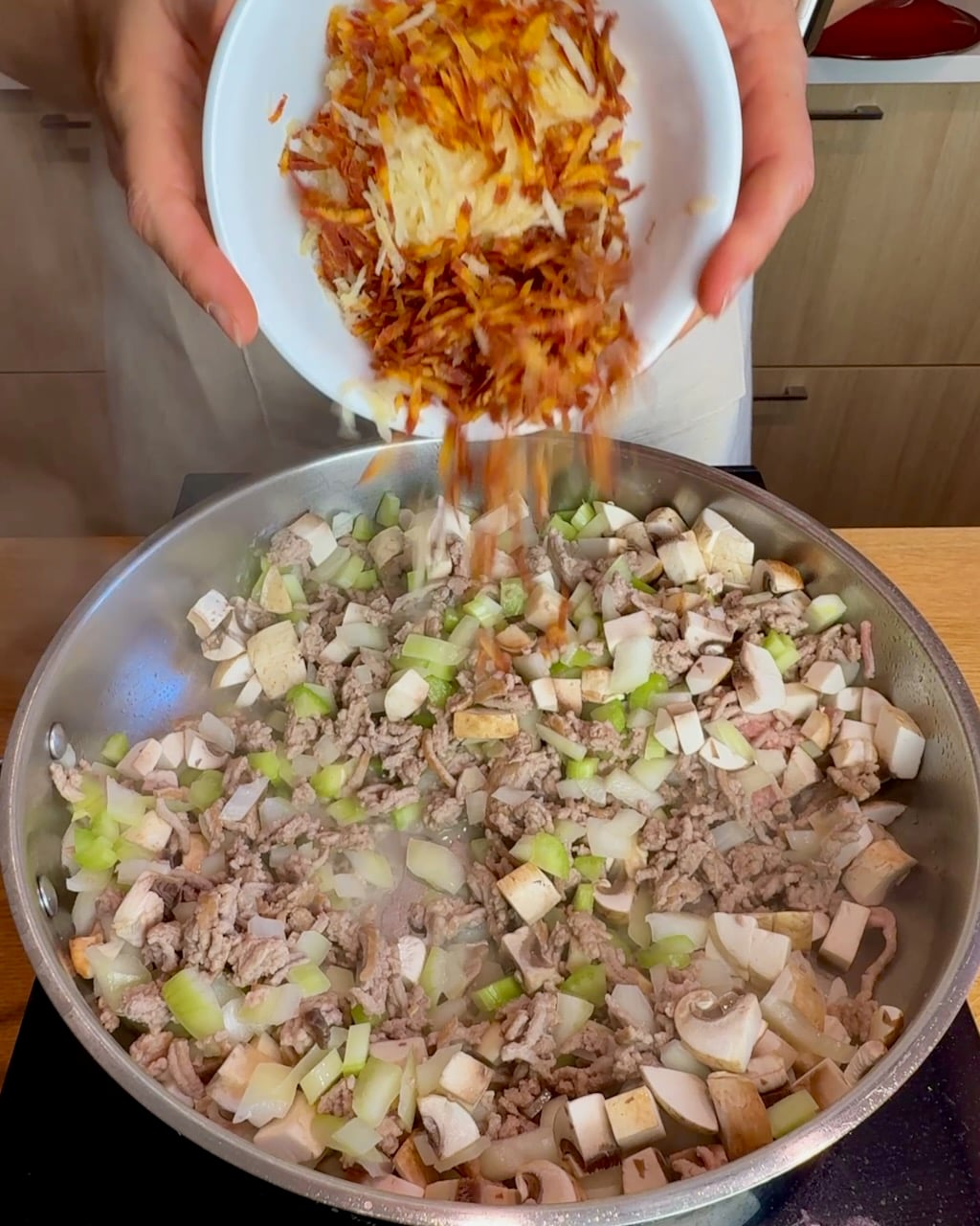 A woman adds grated carrots and other veggies to the ground turkey mixture in the skillet.