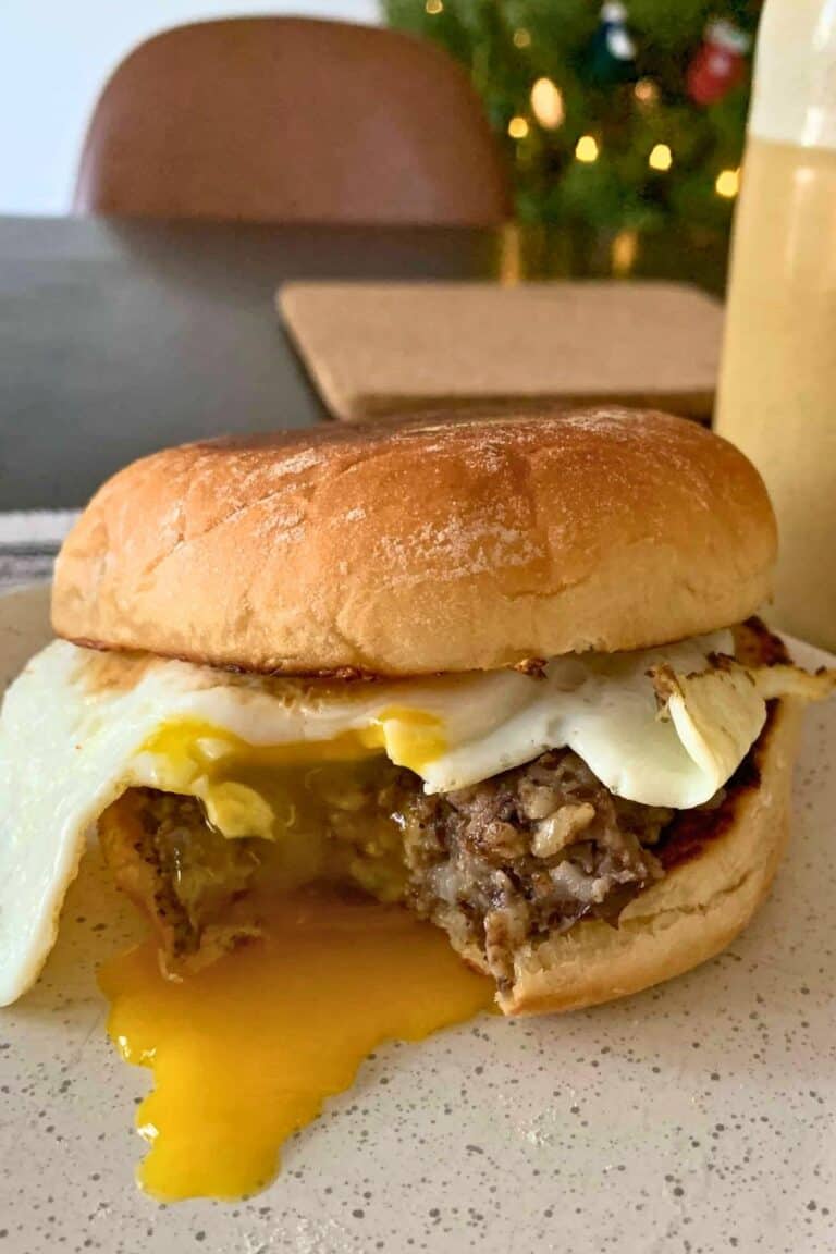 A haggis and fried egg sandwich with a bite taken out of it. The yolk is dripping out onto the speckled off-white plate. There is a cup of coffee and a lit up Christmas tree in the background.