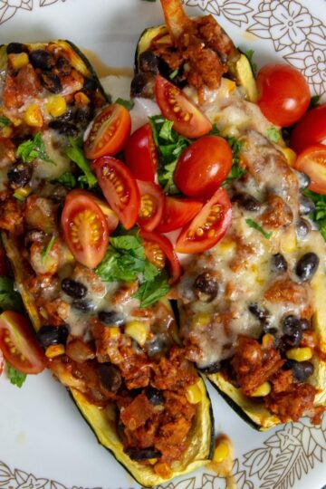 Two turkey taco zucchini boats on a plate with fresh herbs, tomatoes, and a wedge of lime.